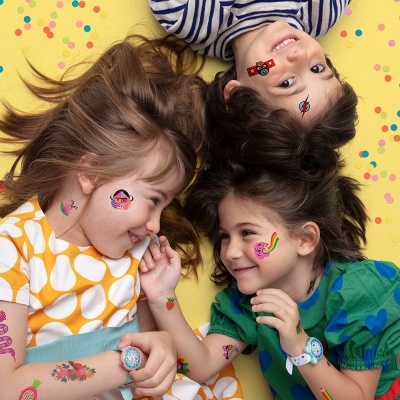 Três crianças com tatuagens temporárias coloridas no rosto e braços, deitadas sobre fundo amarelo com confetes