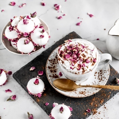 Chávena com bebida cremosa e pétalas de rosa, macarons decorados e colher dourada numa base escura.
