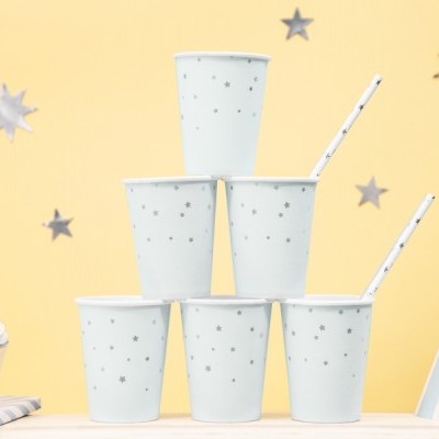 Seis copos de papel brancos com estrelas cinzentas empilhados em pirâmide com canudos sobre mesa clara, fundo amarelo e decoração de festa