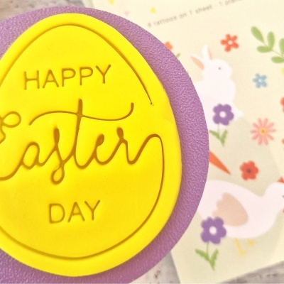 Carimbo amarelo em forma de ovo com texto 'HAPPY Easter DAY' sobre base roxa, com papel decorativo de coelhos e flores ao fundo