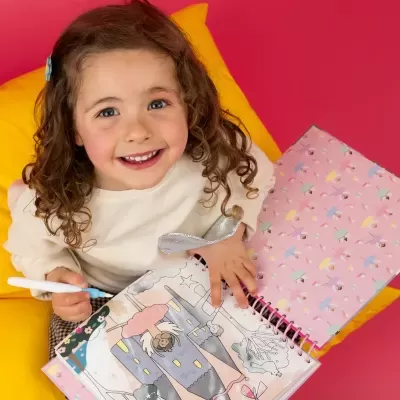 Criança segurando livro de atividades e caneta, sentada em almofada amarela, fundo rosa