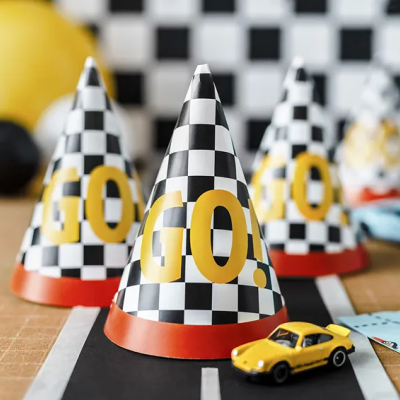 Chapéus de aniversário pretos e brancos de padrão quadriculado com texto GO! amarelo sobre mesa com pista de corrida e carros de brinquedo.