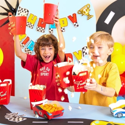 Crianças segurando caixas vermelhas com pipocas e caixas de snacks em forma de carros numa festa decorada com tema de corrida.