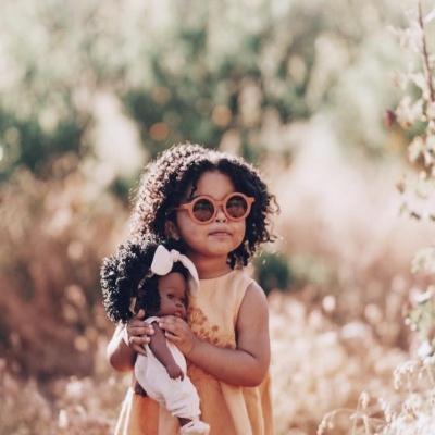 Criança com vestido amarelo e óculos, segurando boneca de pele escura num campo aberto.