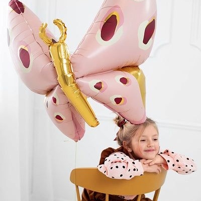Balão borboleta rosa com manchas bordô e dourado segurado por criança numa cadeira castanha clara