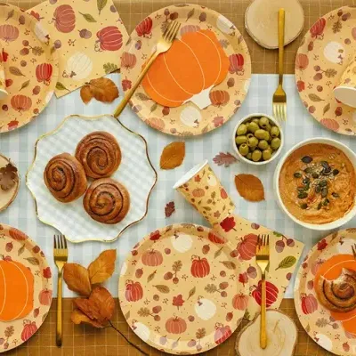 Mesa posta com pratos e copos decorados com abóboras, talheres dourados e comida sobre toalha quadriculada