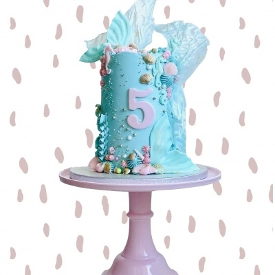 Bolo azul claro decorado com conchas e número 5 em rosa sobre suporte rosa