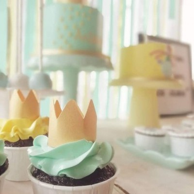 Cupcake com cobertura verde clara e coroa em papel, bolo azul e dourado e bolo amarelo ao fundo