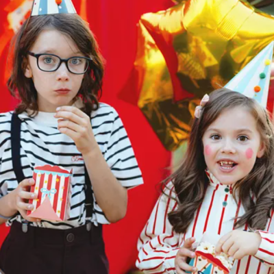 Duas crianças com gorros de festa e caixas de pipocas coloridas junto a balões