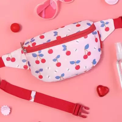 Pochete branca com padrão de cerejas e fecho vermelho sobre fundo rosa com acessórios.