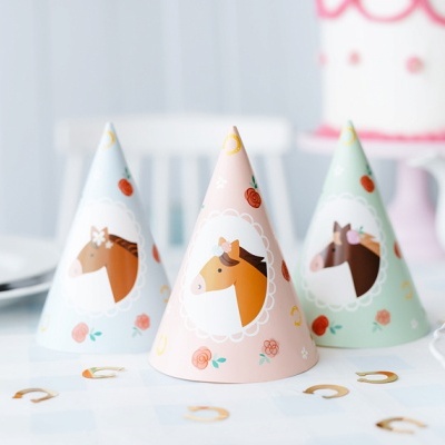 Três chapéus de festa coloridos com estampa de cavalos, sobre uma mesa branca com pratos e bolo ao fundo.