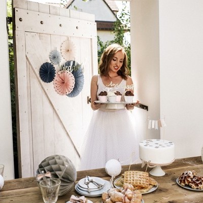 Mulher com vestido branco segurando bandeja com cupcakes à frente de mesa de madeira decorada com doces e elementos de papel coloridos