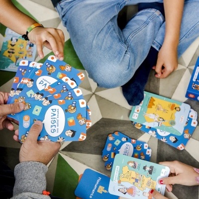 Cartas de jogo educativas com ilustrações coloridas sobre países, vistas em mãos e no chão com padrão geométrico.