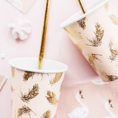 Copos de papel rosa com padrão de penas douradas sobre superfície rosa com cisnes brancos