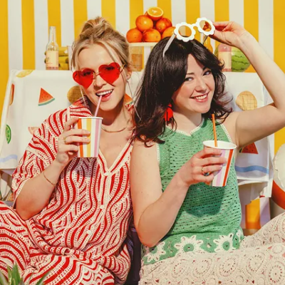 Duas mulheres vestindo roupas coloridas com copos às riscas, sentadas frente a cortinas amarelas e brancas, com frutas e bebidas ao fundo.