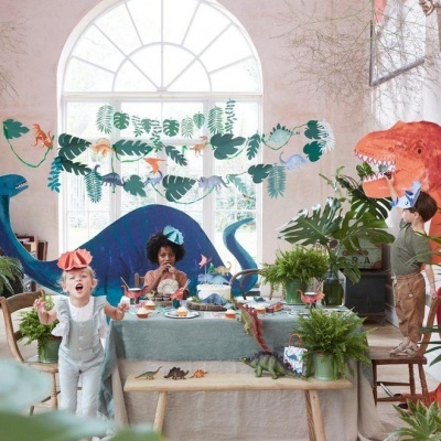 Mesa de festa decorada com tema de dinossauros e crianças vestidas com chapéus de papel coloridos