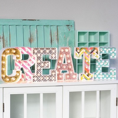 Letras decorativas iluminadas formando CREATE sobre móvel branco com fundo em verde menta envelhecido