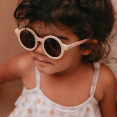 Criança com óculos de sol brancos redondos e camisa de alças branca com padrão xadrez cinza e pequenos desenhos cor de laranja