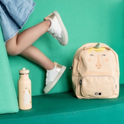 Mochila de unicórnio bege e garrafa na mesma cor junto de criança com sapatilhas e saia azul sentada num sofá verde.