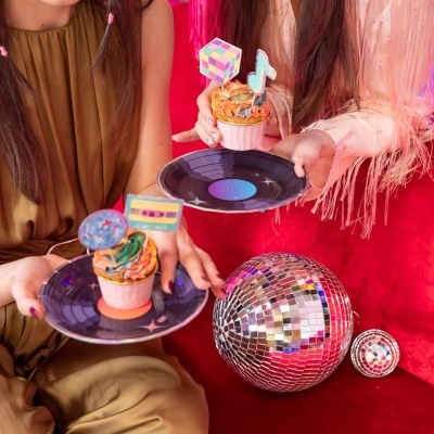 Dois cupcakes coloridos decorados com toppers em pratos pretos sobre fundo vermelho com bolas de discoteca