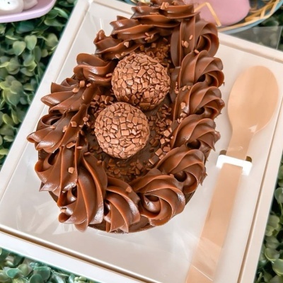 Ovo de Páscoa de chocolate decorado com trufas e pedaços de chocolate em embalagem branca com espátula rosa.