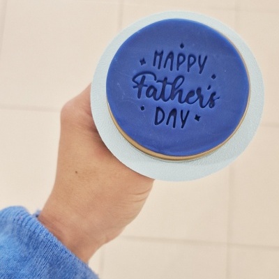 Produto redondo azul-escuro com texto 'HAPPY Father's DAY' segurado por mão com manga azul