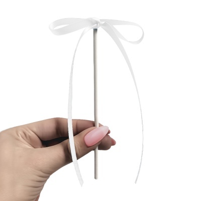 Palito branco com laço branco segurado por mão feminina com unhas cor de rosa