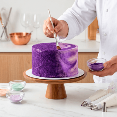 Bolo redondo cor roxa a ser pintado com pincel, em suporte de madeira, com utensílios de confeitaria ao lado