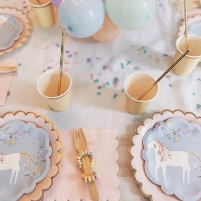 Mesa de festa infantil com pratos de papel com unicórnios, copos amarelos, talheres de madeira com braceletes de doces e balões azuis
