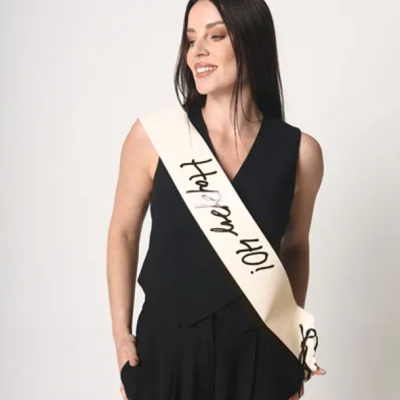 Mulher com roupa preta e faixa branca '¡Oh baby!' em fundo branco