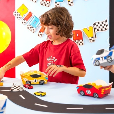 Criança a brincar com carrinhos de corrida e caixas de batatas fritas em forma de carro numa mesa com decoração de estrada