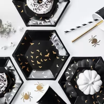 Conjunto de pratos e copos pretos com estampas douradas, donuts brancos e abóbora branca numa mesa branca