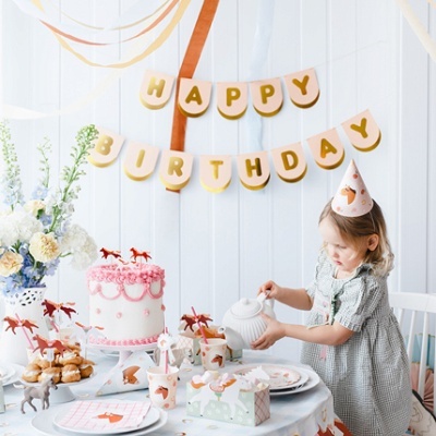 Decoração de festa de aniversário com bolo, copos e toalha com padrão de cavalinhos e menina a servir chá.