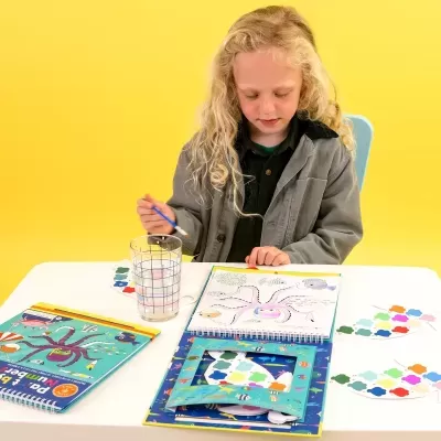 Criança loira a pintar livro de atividades com tema marinho numa mesa branca com fundo amarelo