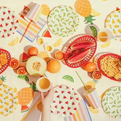 Mesa posta com pratos de fruta, cestas vermelhas com snacks e copos coloridos sobre toalha com estampa de frutas.