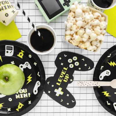 Mesa com artigos de festa com tema videojogos, incluindo pratos, guardanapos, copos, maçã, pipocas e café.