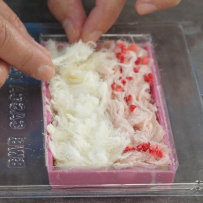 Barra de sabonete rosa claro e branco com pedaços vermelhos dentro de embalagem transparente.