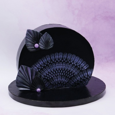 Bolo decorado preto com padrão azul e folhas decorativas com pérolas roxas em fundo lilás