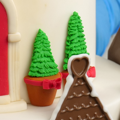 Detalhe de bolo decorado com pinheiros verdes em vasos castanhos e casinha de chocolate com glacê branco