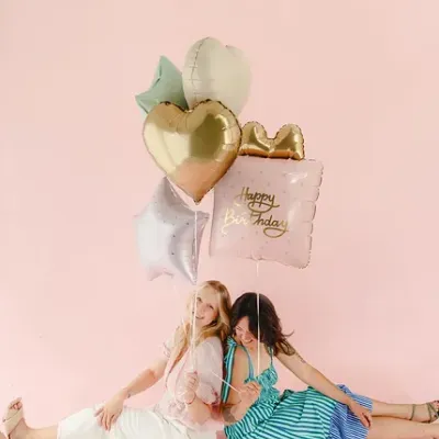 Duas mulheres sentadas com balões coloridos de aniversário em fundo rosa