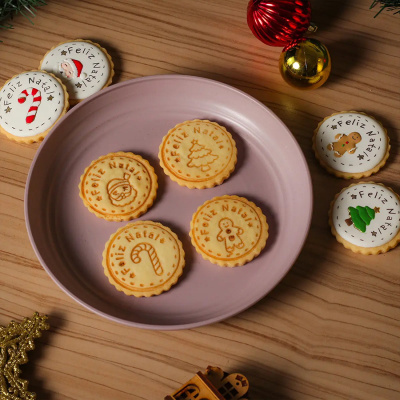 Bolachas redondas de Natal com mensagens e desenhos decorativos em prato rosa com enfeites natalícios numa mesa de madeira.