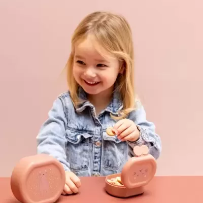 Menina loira a segurar bolacha, com duas caixas rosa com desenhos de gatos, numa mesa rosa.