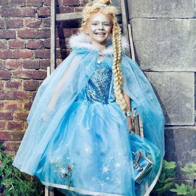 Menina com vestido e capa azul, penteado com trança, segurando bolsa, junto a escada de madeira