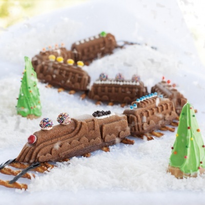 Comboio de chocolate com confeitos coloridos e árvores de Natal verdes em cenário de neve sintética.