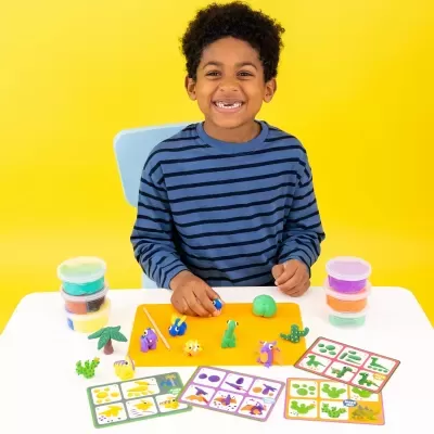 Criança sorridente com camisola às riscas azuis, massinha colorida e cartões de instruções à mesa.
