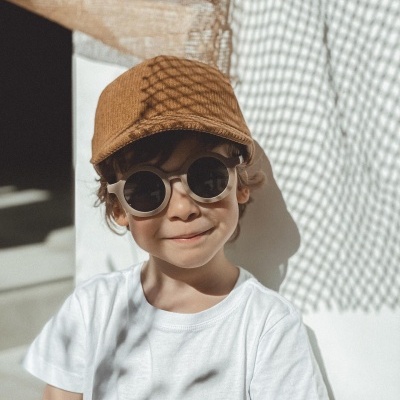 Criança com boné castanho e óculos de sol redondos, t-shirt branca, contra uma parede branca com sombra de rede quadriculada