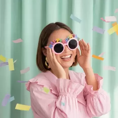 Óculos HAPPY BIRTHDAY rosa usados por mulher em fundo com cortinas verdes