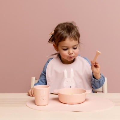 Criança pequena com babete rosa sentada com conjunto de refeição rosa numa mesa com fundo rosa.