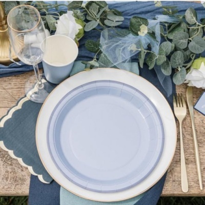 Mesa posta com prato branco e azul, talheres dourados, copos de plástico, flores e folhagem sobre toalha azul escura