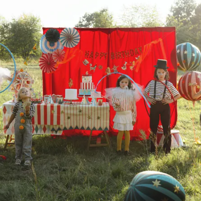 Crianças vestidas de circo em cenário ao ar livre com cortinas vermelhas e balões listrados